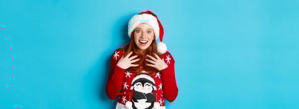 Happy holidays and Christmas concept. Surprised redhead girl receiving unexpected gift, gasping amazed and staring with plesant disbelief, standing in santa hat against blue background photo