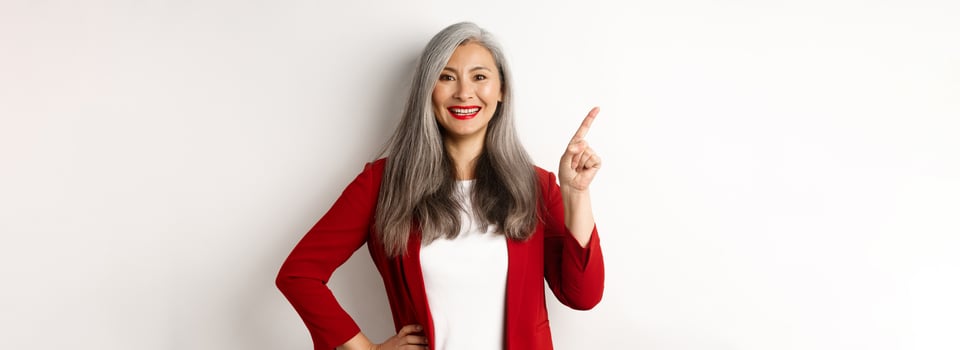 Happy senior office lady pointing finger at upper left corner, showing company logo and smiling at camera, standing over white background photo