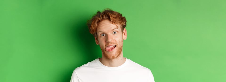 Emotions and fashion concept. Close up of funny redhead man showing silly faces, sticking tongue and staring at camera, standing over green background photo