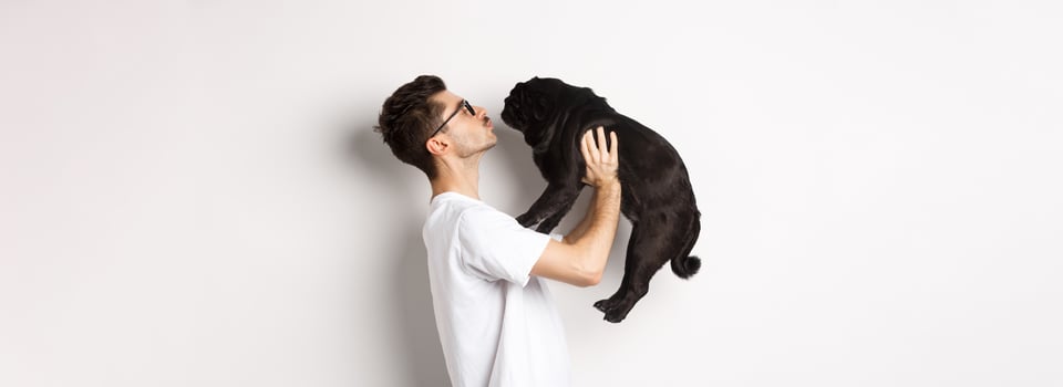Profile of handsome young man kissing small cute dog face. Hipster guy loving his pug, standing over white background photo
