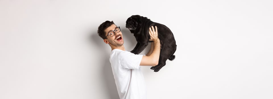 Image of handsome young man loving his pug. Dog owner holding puppy and smiling happy at camera, standing over white background photo