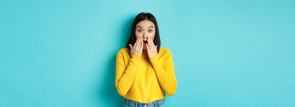 Portrait of impressed korean girl saying wow, gasping and staring amazed at camera, standing in yellow pullover over blue background photo
