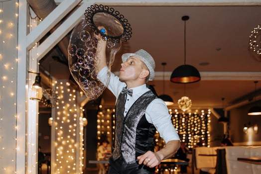A man in a black vest and hat blowing bubbles. The bubbles are in a clear plastic container photo