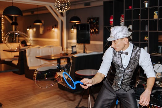 A man in a black vest and hat is blowing bubbles. The bubbles are floating in the air and the man is holding a blue bubble wand. The scene is set in a restaurant with tables and chairs photo