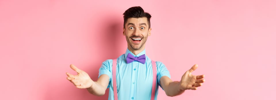 Happy handsome man in bow-tie stretch out hands in warm welcome, inviting or greeting you, receiving gift, standing on pink background photo