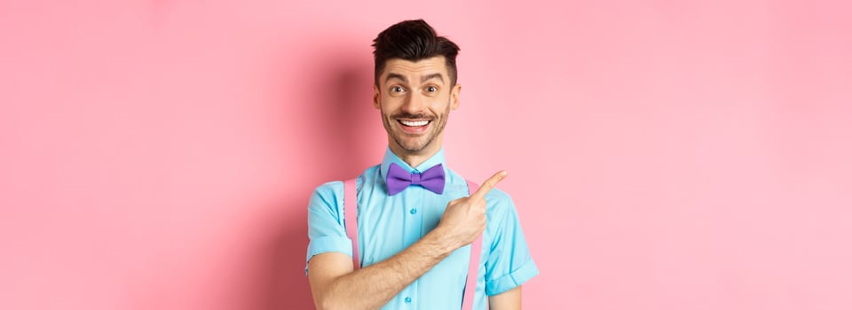 Portrait of handsome happy man showing logo, pointing left and smiling at camera, recommending shop offer, standing on pink background photo