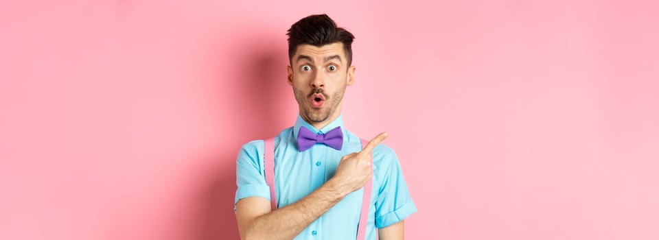 Check it out. Amazed caucasian man with moustache, wearing bow-tie, saying wow and pointing left, showing advertisement, standing on pink background photo