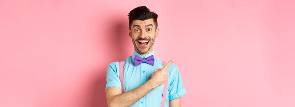 Happy young man checking out promo offer, smiling amazed and pointing finger left at copy space, standing near logo on pink background photo