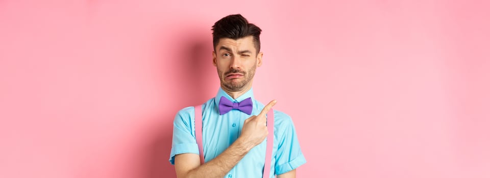 Childish young man acting sad and fake crying while pointing finger upper left corner, complaining on something, standing over pink background photo