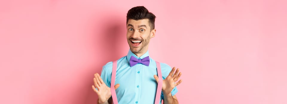 Cheerful guy in bow-tie, showing his suspenders and smiling happy, feeling upbeat, standing excited over pink background photo