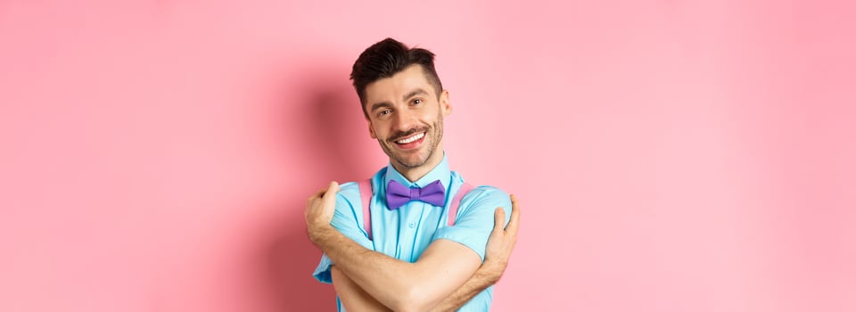 Cheerful young man smiling and hugging himself, wrap hands around own body and looking happy, standing over pink background photo