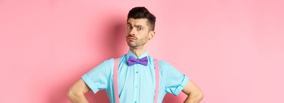 Image of young man with funny moustache and bow-tie standing in confident pose, staring with disbelief and doubts at camera, posing over pink background photo