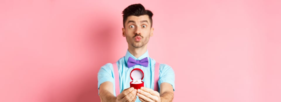 Valentines day. Funny young man pucker lips for kiss and showing engagement ring, making proposal, say marry me to lover, standing over pink background photo