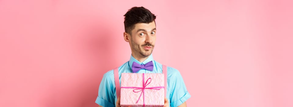 Holidays and celebration concept. Funny guy with moustache showing cute gift box wrapped in festive paper, looking at camera, presenting you, standing on pink background photo