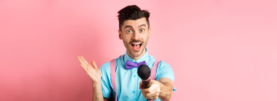 Cheerful young man, show host in bow-tie giving you microphone and smiling, entertain people on festive celebration events, performing at holidays, standing on pink background photo