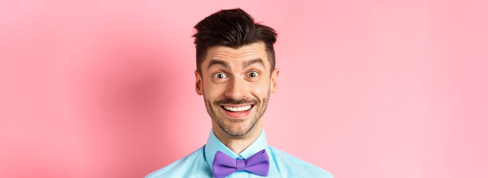 Close-up of cheerful caucasian man smiling happy, looking at something interesting, standing in bow-tie over pink background photo