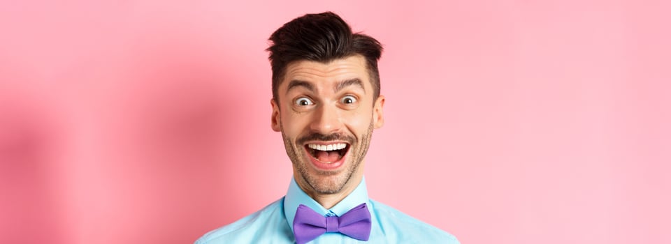 Close-up of surprised and happy young guy, open mouth and smiling amazed, checking out promo offer, standing in bow-tie over pink background photo