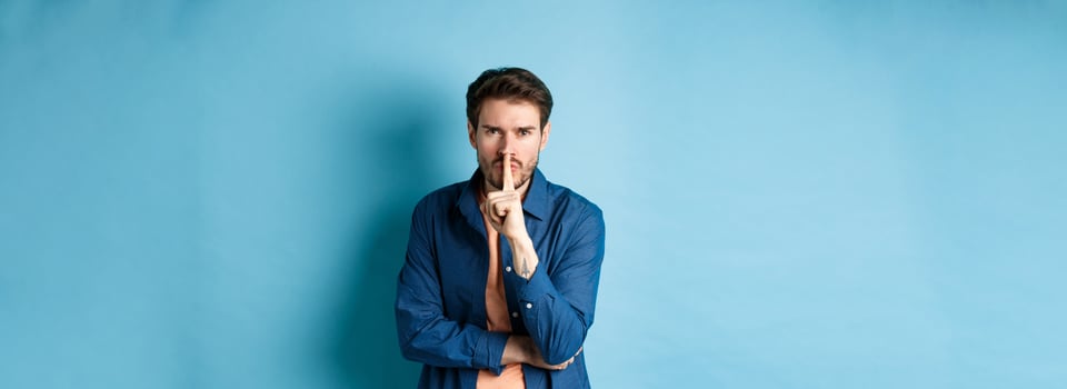 Angry man shushing, press finger to lips and frowning, tell to be quiet, show taboo gesture, standing on blue background photo