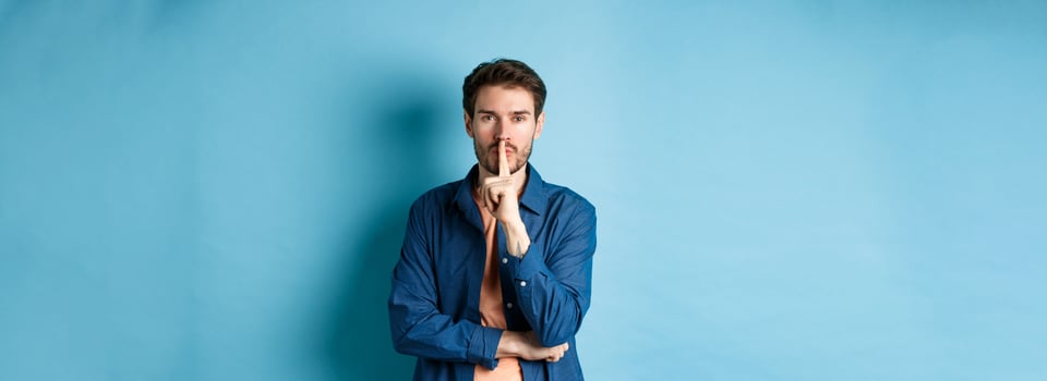 Modern guy shushing at camera, press finger to lips and telling a secret, making taboo sign, standing on blue background photo