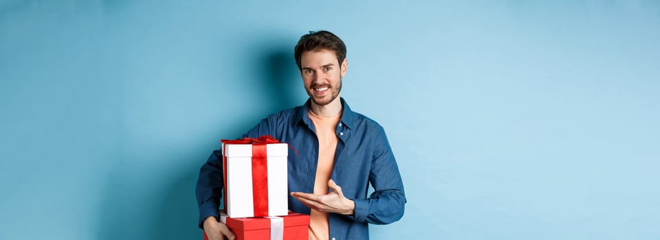 Young man in casual clothes buying romantic gifts on Valentines day, pointing at presents boxes and smiling, standing over blue background photo