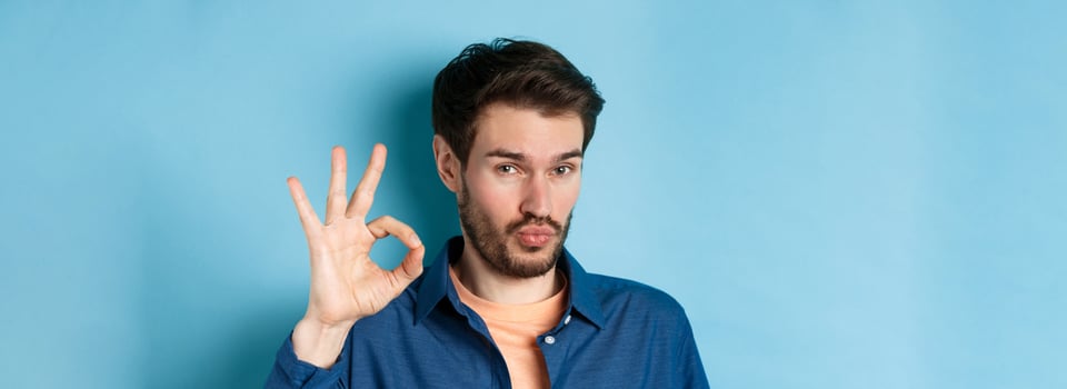Image of silly young man pucker lips and show okay gesture, praise good thing, compliment excellent choice, standing on blue background photo