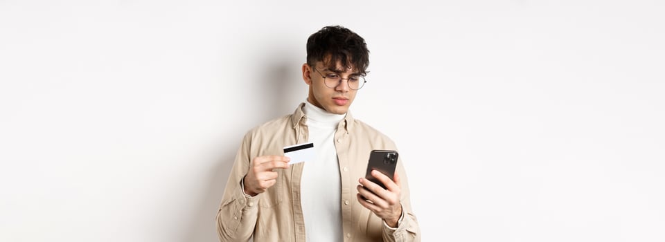 Handsome young man in glasses making purchase on phone, shopping online, holding plastic credit card and smartphone, standing on white background photo