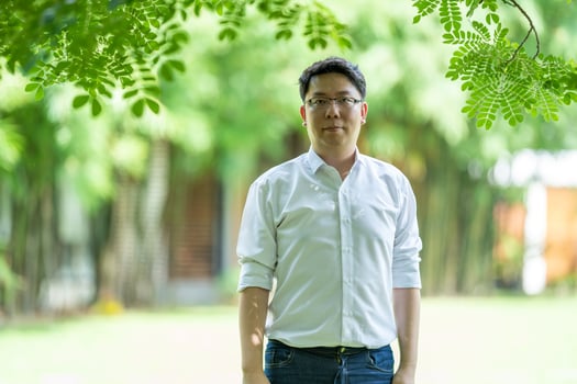 Looking through the garden is a trusty Asian man with a white shirt, black pants, and glasses post under the big thee with the green garden blur background. photo