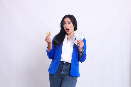 beautiful adult office girl wearing blue formal suit standing surprised to camera hand holding dollar money and bitcoin isolated white background. for transaction content, investment, business photo