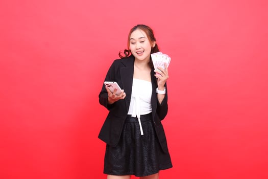 expression of young indonesia career cheerful woman wearing black formal suit, hand holding rupiah money and cell phone up, for technology, business, advertising, financial content photo