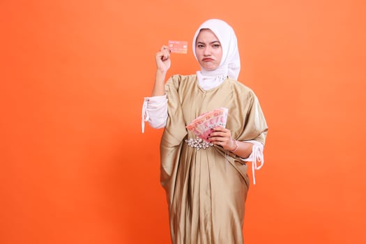 young asian woman frowning holding rupiah money down and member card, debit credit card wearing kaftan Muslim dress with hijab, isolated on orange background. Fashion concept, publication, promotion photo