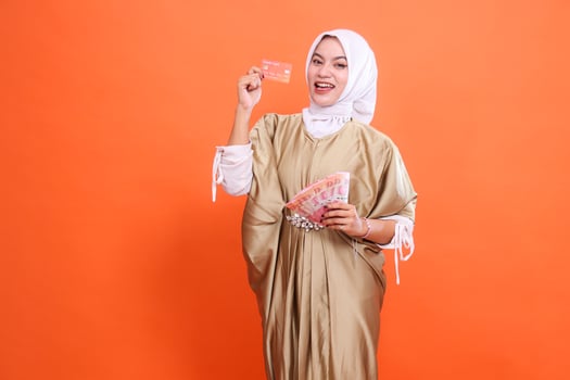 Cheerful young Asian woman holding rupiah money down and member card, debit credit card wearing Muslim dress kaftan with hijab, isolated on orange background. Fashion concept, publication, promotion photo