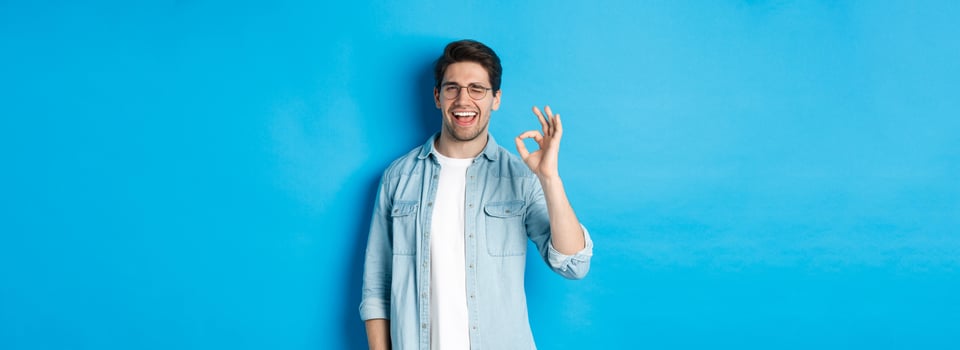 Confident smiling man in glasses showing ok sign, winking to guarantee or recommend something, blue background photo