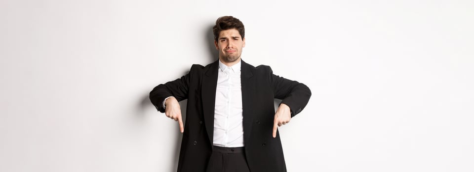 Portrait of skeptical and unamused bearded man, pointing fingers down with disappointed face, standing in suit and complaining photo