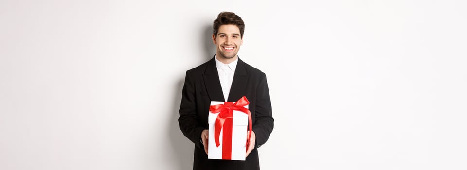 Concept of christmas holidays, celebration and lifestyle. Handsome man in black suit, have romantic present, holding gift in a box and smiling, standing against white background photo