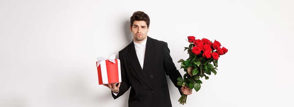 Concept of holidays, relationship and celebration. Handsome and confident man in black suit, going on a date, holding bouquet of roses and present, standing against white background photo