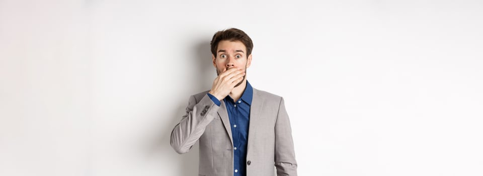Shocked boyfriend in suit gasping, covering mouth with hand and pop eyes at camera startled, standing on white background photo