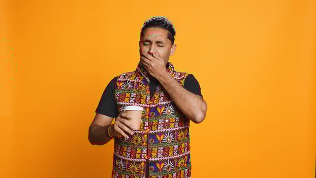 Tired indian man sipping fresh coffee from disposable paper cup early in the morning to wake up and be energized. Person drinking hot beverage from recycled takeaway cup, studio background, camera B photo