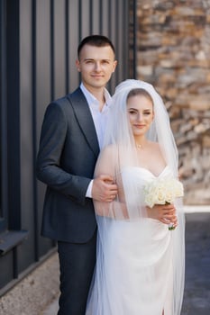 A bride and groom are posing for a picture in front of a building. The bride is holding a bouquet of flowers and the groom is wearing a suit. Scene is happy and celebratory photo