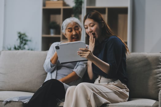 Happy grownup daughter showing content on tablet to mature mother. Two family generations women with digital computer gadget resting on couch together, watching videos, making call photo