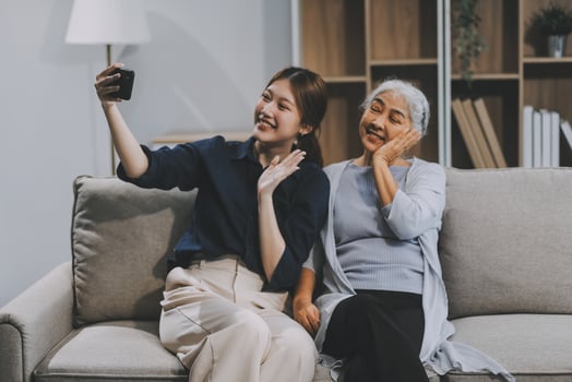 Cheerful mother and pretty teenager kid relaxing at cozy home, using mobile phone for Internet communication, resting on sofa with digital gadget, enjoying leisure, looking at camera photo