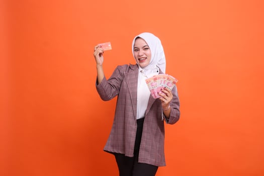 Cheerful 20s young Asian girl wearing hijab office wear holding debit credit card and money to the side isolated on plain pastel orange background studio portrait photo