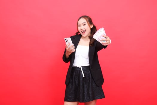 gesture of young asian career cheerful woman wearing black formal suit, hands holding rupiah money and smartphone in front, for technology, business, advertising, financial content photo