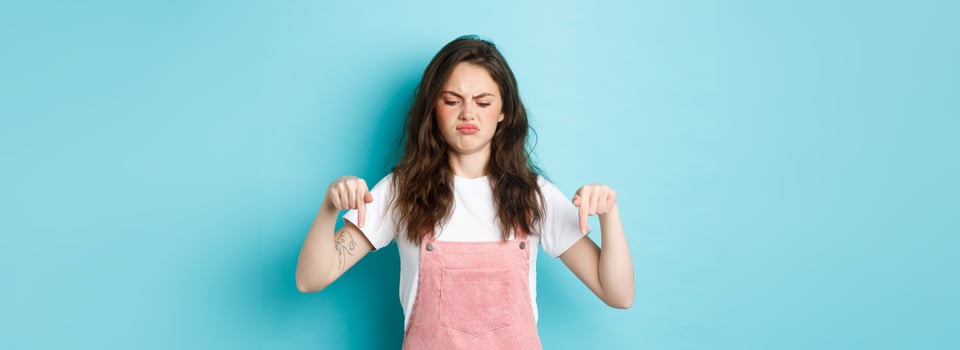 Sounds bad. Displeased grumpy brunette girl frowning and sulking moody, pointing fingers down and look disappointed, standing against blue background photo