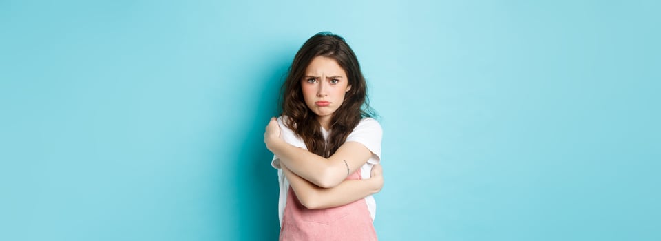 Sad and gloomy young woman pouting, comforting herself with cuddles, hugging her body and looking upset about something unfair, standing against blue background photo