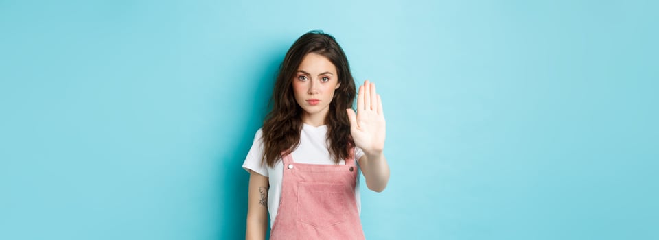 Young brunette woman with curly hairstyle, raising hand in block gesture, say stop or no, refuse bad offer, rejecting something, standing against blue background photo