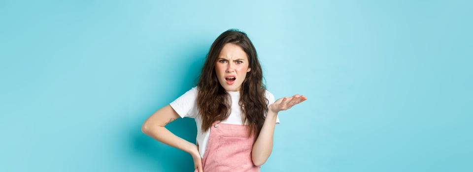 Image of confused and annoyed young woman raising hand and pointing at something terrible, staring puzzled at camera with pissed-off face, standing over blue background photo