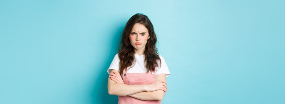 Portrait of angry and upset sulking girl, pouting about unfair situation, frowning and staring at camera, cross arms on chest defensive, standing over blue background photo