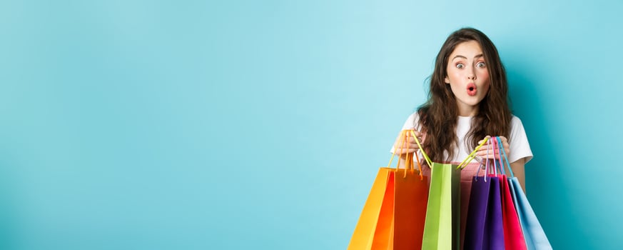 Amazed young woman shopaholic, holding colorful shopping bags and look amused at next shop, buying things in store, standing over blue background photo