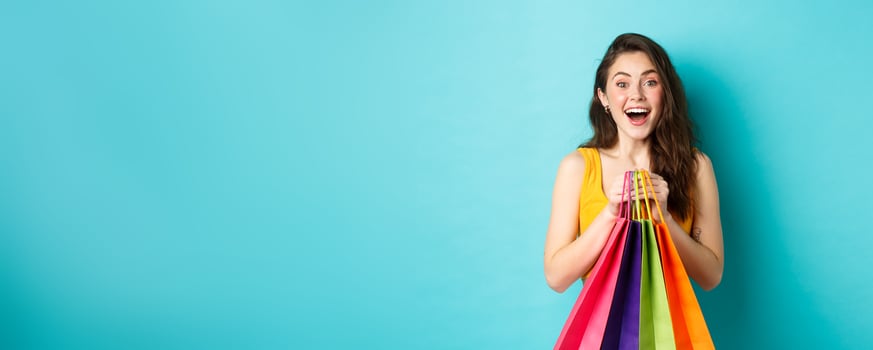 Portrait of surprised young woman look amazed at camera, holding shopping bags, see discounts in store, standing over blue background photo