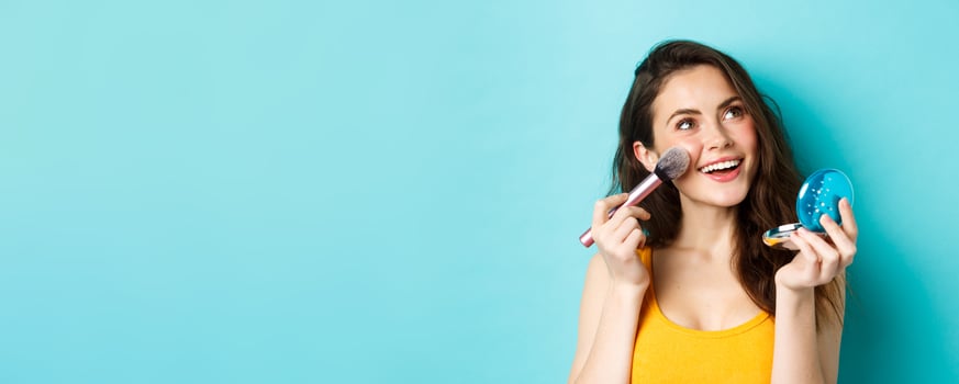 Beauty. Young glamour girl applying make up with pocket mirror and brush, smiling and gazing up at logo, standing against blue background photo